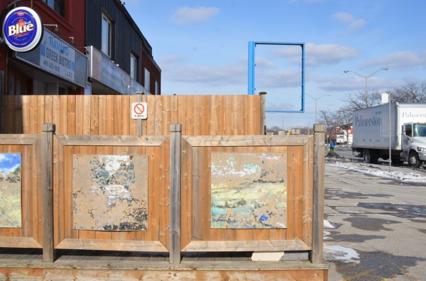 wooden fence around a patio, with two old paintings that are faded and peeled so you can't tell what they were pictures of, in the background, an empty blue metal frame that once held a sign for a store 