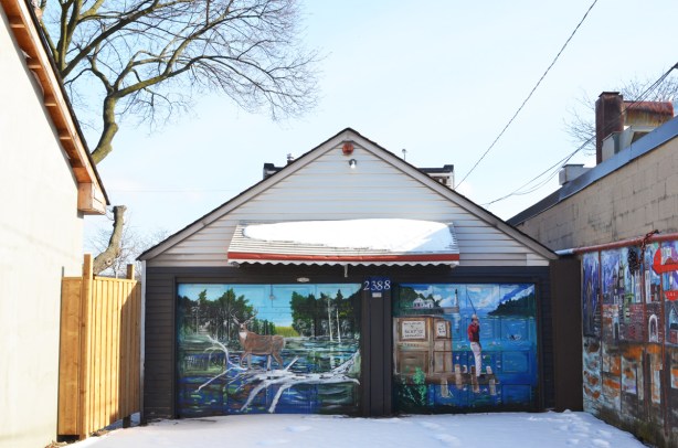 two garage doors side by side in an alley with murals painted on them, a deer with antlers on the left and a man fishing in a river on the right 