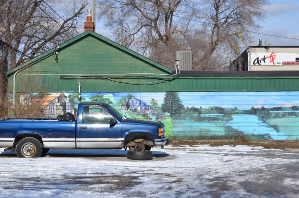 a blue pickup truck with one tire missing, parked in a vacant lot, in front of a farmyard scene mural with fields and a pond