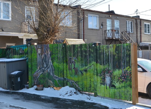 a woodlands theme mural on a wood fence between two properties in a lane, a tree trunk in the mural matches the large tree behind the fence 
