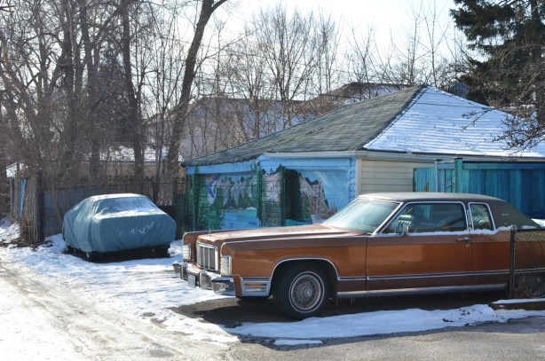 an old rust coloured Lincoln Town Car parked in an alley, garage doors behind it are covered in murals by bc johnson 
