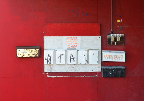 a red wall in front of a building, mailboxes on it, 8 mailboxes, also two buzzers under a sign that says Supt Bob 