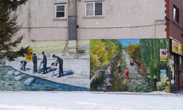 two historical murals on the side of a building, one is boys ice fishing and the other is people swimming in the creek 