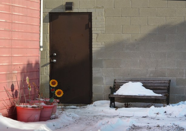 brown metal door on concrete block wall, pink planters with fake sunflowers in them, a bench with snow on it beside the door too 