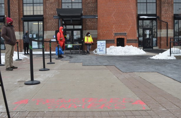 people at skating rink, outdoors. one is secutiy and others are waiting their turn to use the ice. pink letters on sidewalk that denote place for those with reservations to line up 