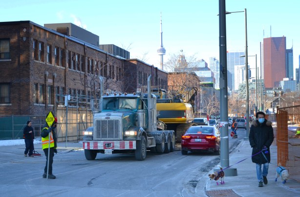 flat bed truck carrying machinery, and little red car on street, man holding slow stop sign by construction site , man on sidewalk walking two small dogs
