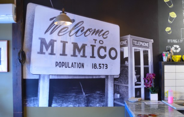 large photograph on a coffee shop wall of a welcome to mimico sign beside an old phone booth with someone inside it 