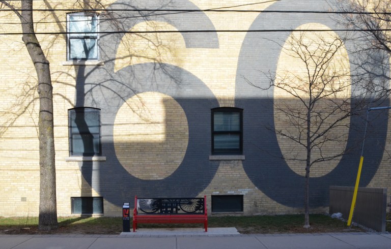 black and red metal bench in front of a two storey beige brick wall with a large number 60 painted in black on it 