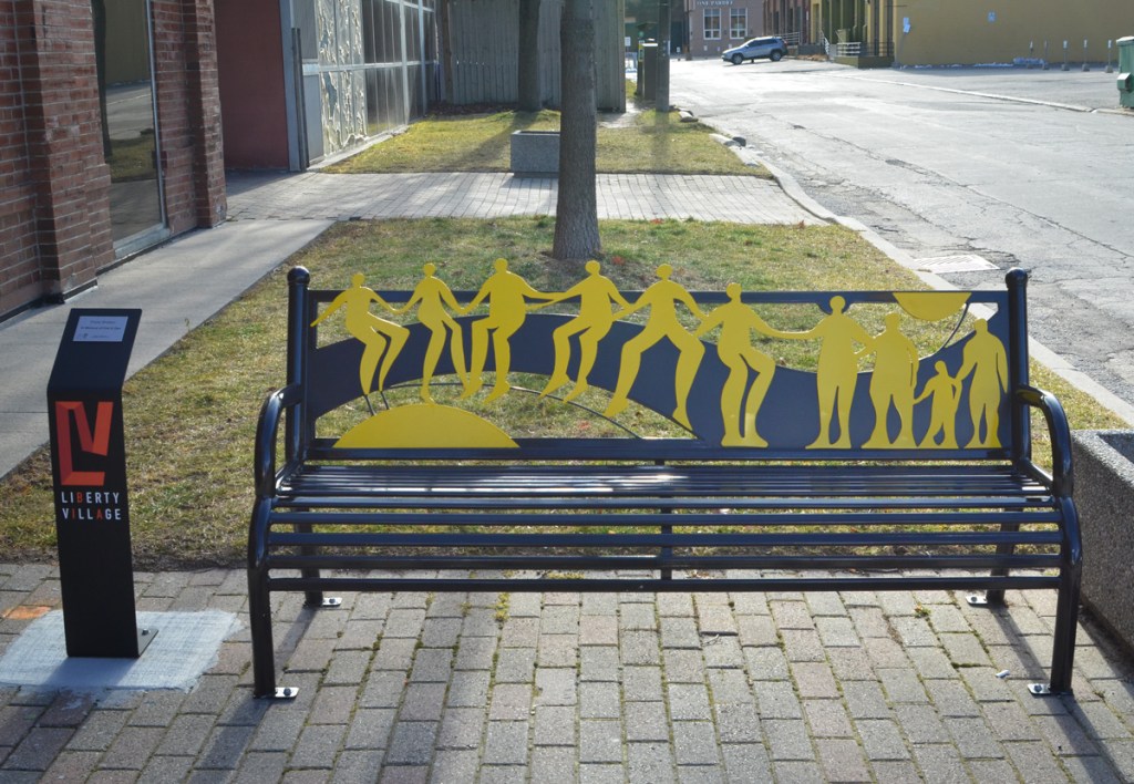A black metal bench between the sidewalk and street, the back of the bench has yellow figures all holding hands