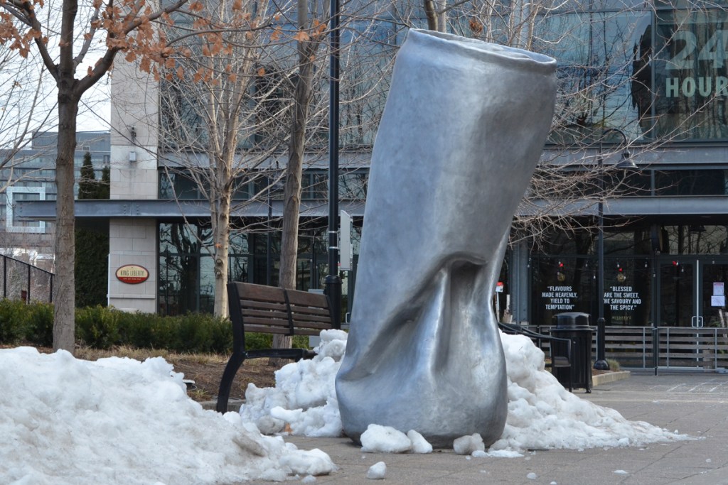large sculpture of a crushed metal can, grey, beside a pile of snow on a pedestrian walkway in Liberty Village 