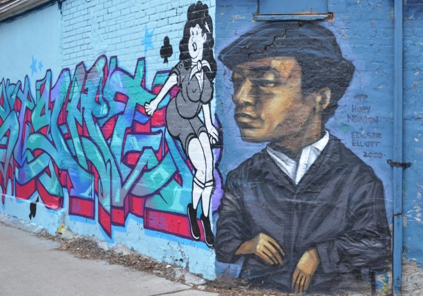 Huey Newton mural by elicser on the end of a wall, a mural beside it with a buxom woman in tight clothes