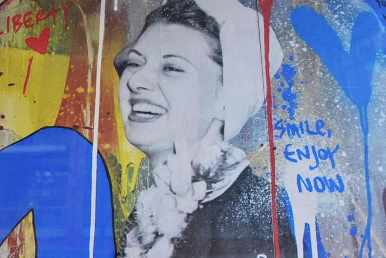 black and white picture of a woman in a white hat and white fluffy scarf on hoardings with blotches of yellow and blue, also blue writing that says smile enjoy now 