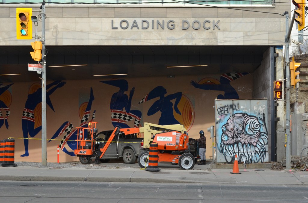 grominator graffiti on a metal box beside sidewalk, a cherry picker lift machine parked beside it, under an overhang that says loading dock.  Mural of black people dancing on the wall behind the lifter. 