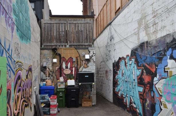 mural of cartoon character Yosemite Sam, on a wall at the end of a driveway, with trash cans and other stuff in front of it 
