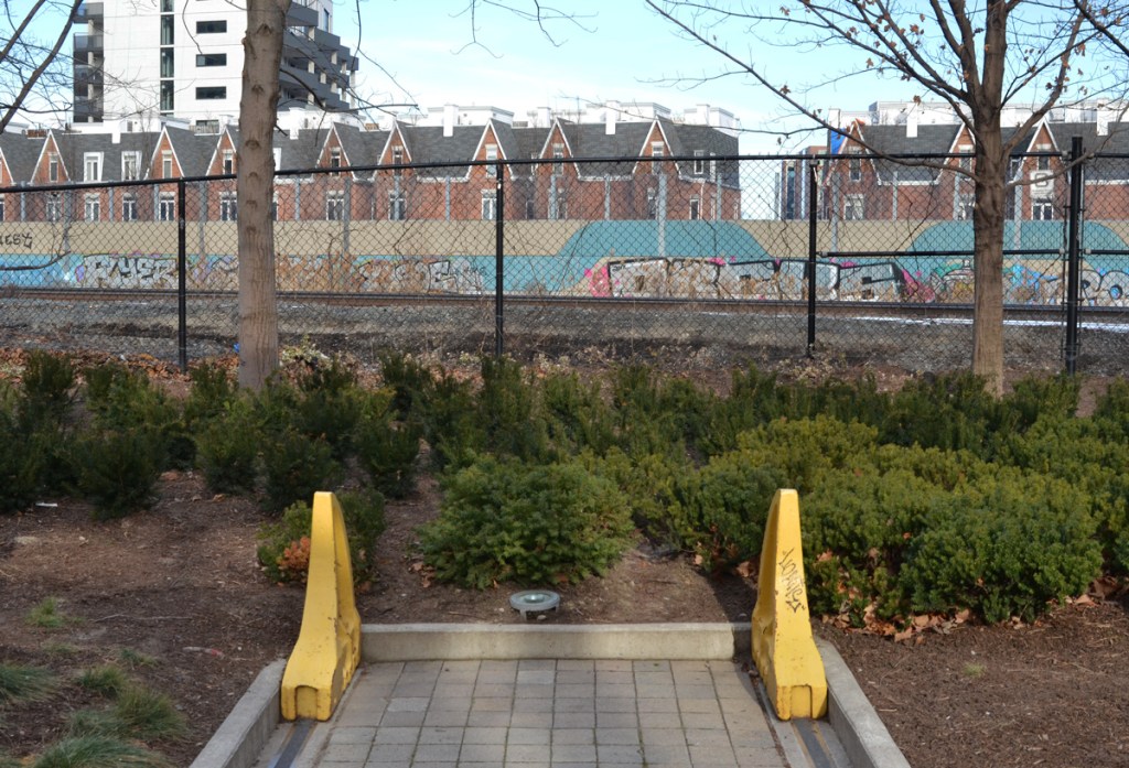 two yellow metal bumpers at the end of the railway tracks, left over from a real railway line, ends in park by a fence along another, real, train tracks, graffiti on the walls beyond the tracks, houses beyond that  