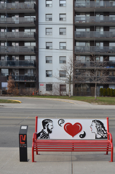 an apartment building across the street, a bench on the sidewalk in the foreground.  The bench is red with a picture on the back of a man and woman yelling at each other and a red heart in the middle 