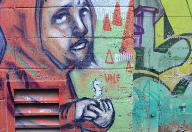 part of elicser mural of a man with a small beard in red hoodie, holding a small unicorn in his hand 