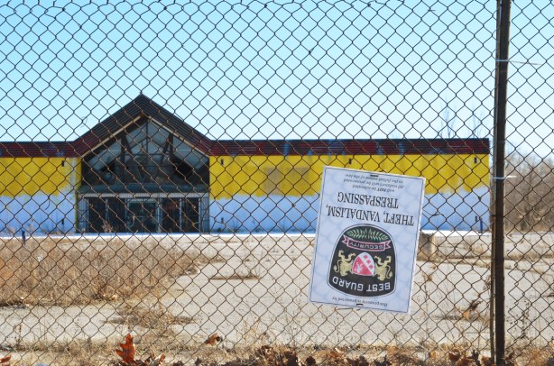 chainlink fence around an empty and abandoned store with yellow and white front, large vacant parking lot in front, a security sign is upside down on the fence 
