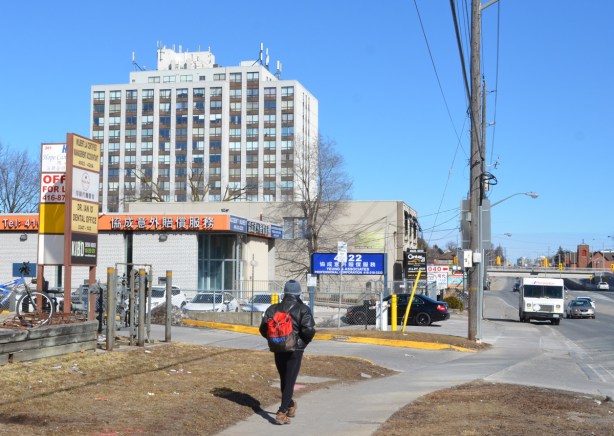 a man with a red backpack walks along the sidewalk in front of some small businesses, a condo building in the background 