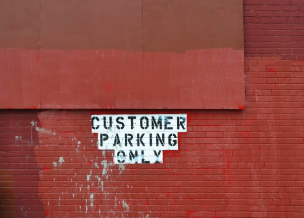 brick wall with a wood sign, all painted over in red, in the middle is black paint on white words that say customer parking only 