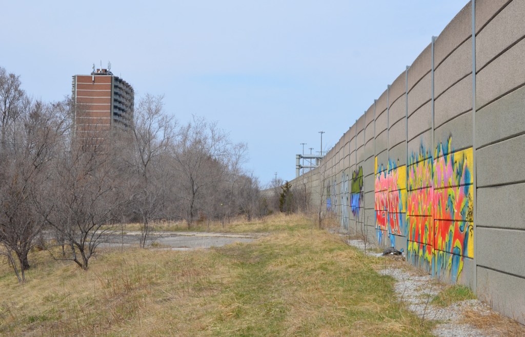 text graffiti on a concrete wall, beside green space and the dead end of an abandoned onramp for the Don Valley Parkway, red brick apartment building, small trees, 