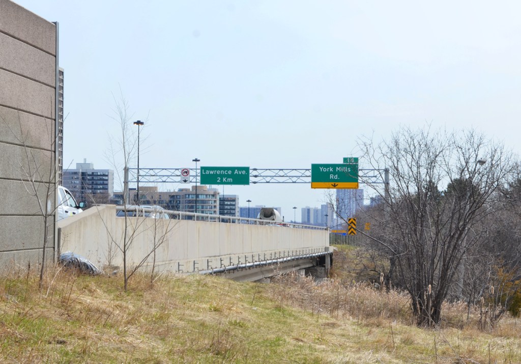 up on a hill, level with a DVP overpass over York Mills Road, concrete barrier at side of parkway, signs for exit ramp to York Mills, also sign saying Lawrence Ave in 2 km, 