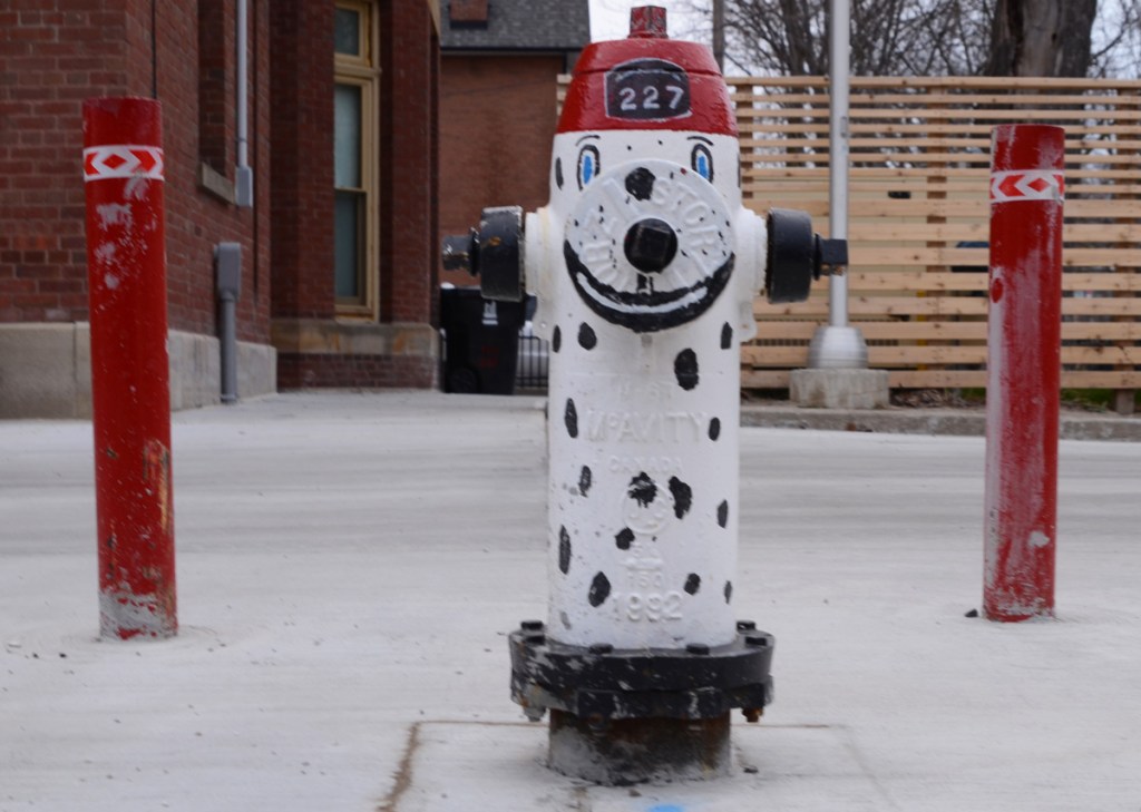 fire hydrant in front of a fire station 227 painted like a dalmation dog, white with black spots, a red hat with number 227 on its head, and a big smile on its face
