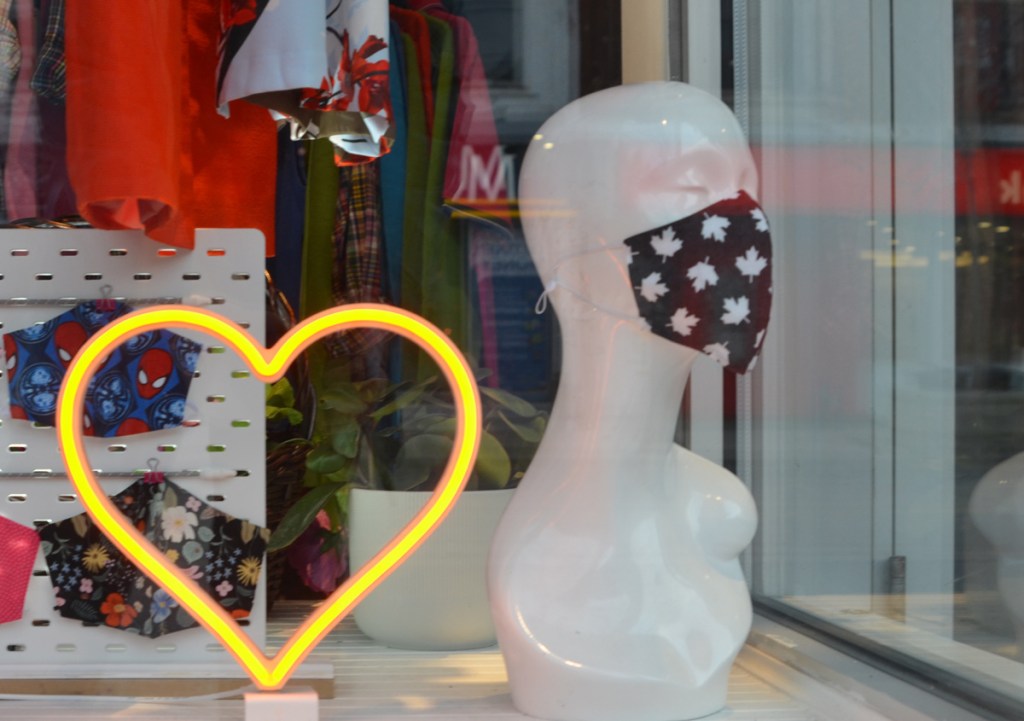 store window with a yellow LED light heart, a white head and shoulders mannequin with closed eyes and wearing a black covid mask with white maple leafs on it.  