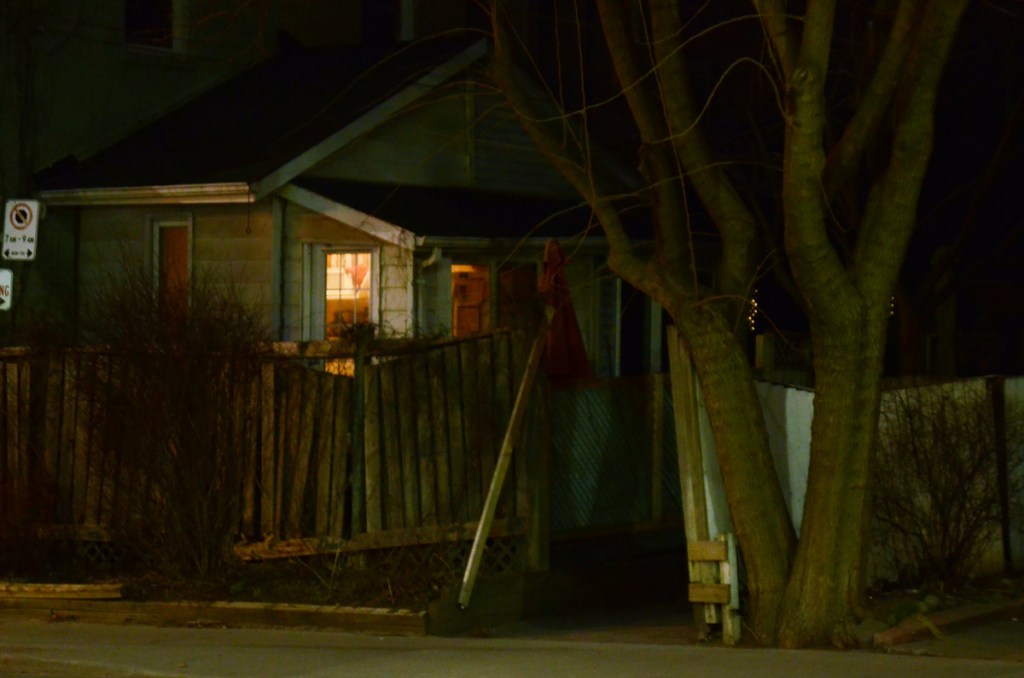 back of a small house at night, gate open, light on in back room, tree by sidewalk, 