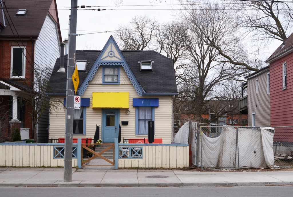 small white house with blue trim and a yellow awning over the front door, white fence around the front yard, empty lot beside it with white tarp covering chainlink fence around the yard