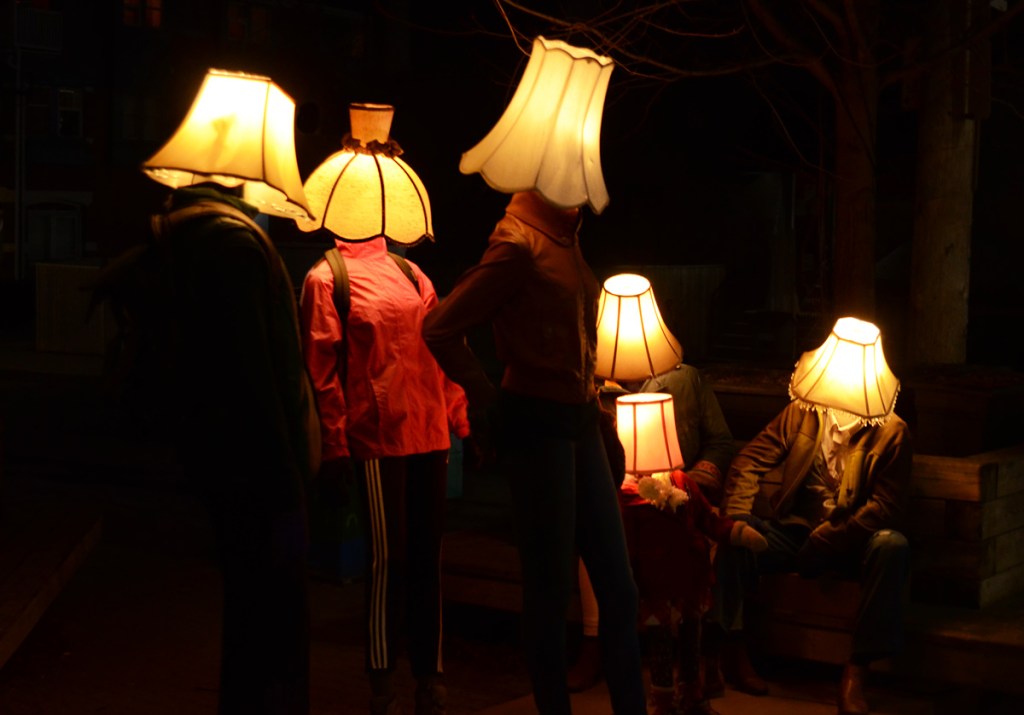 Luminosity art installation of mannequins with lampshades for heads.  night time, so dark outside, lights turned on inside shades