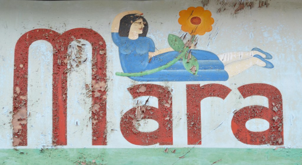 old sign above a store doorway, the word Mara with a woman in a blue dress lying across the last 3 letters, holding a big orange and red flower 