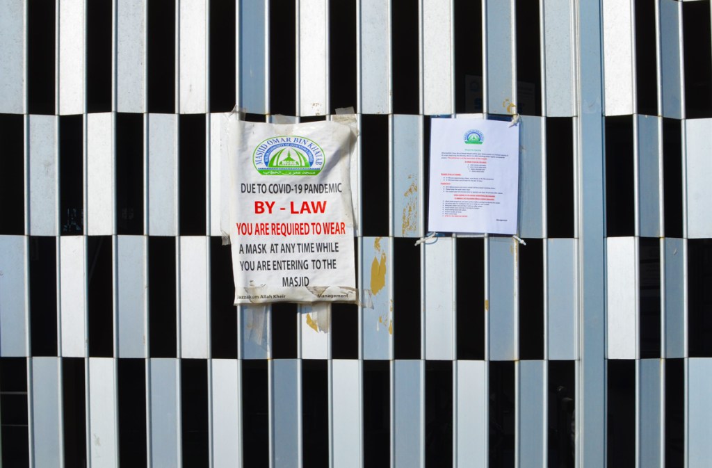 metal gate over entrance to masjid with a sign on it was due to covid pandemic, bylaw you are required to wear a mask at anytime while you are entering the masjid 