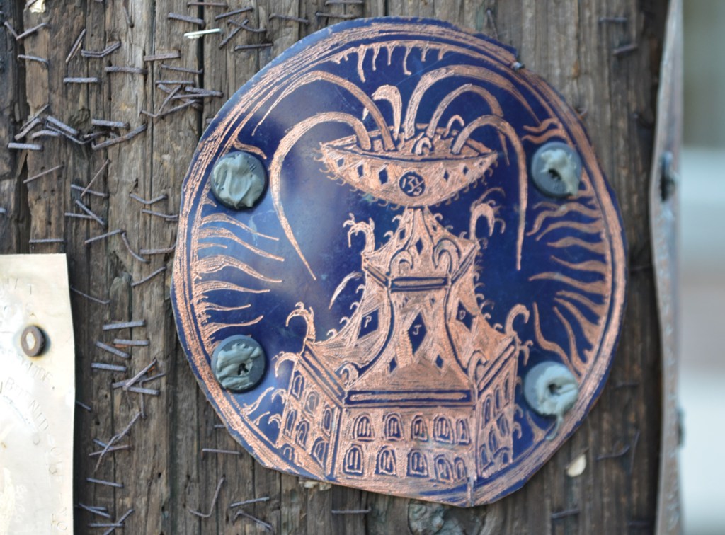 round metal engraving on a small circle, screwed to a wood utility pole. Design is like a fountain on the top of a building 13 1/2