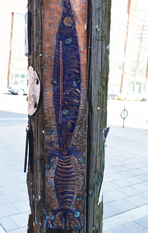 a long narrow, vertical, engraved metal graffiti on a wood pole. Latin words are written around a fish like creature shape with designs inside