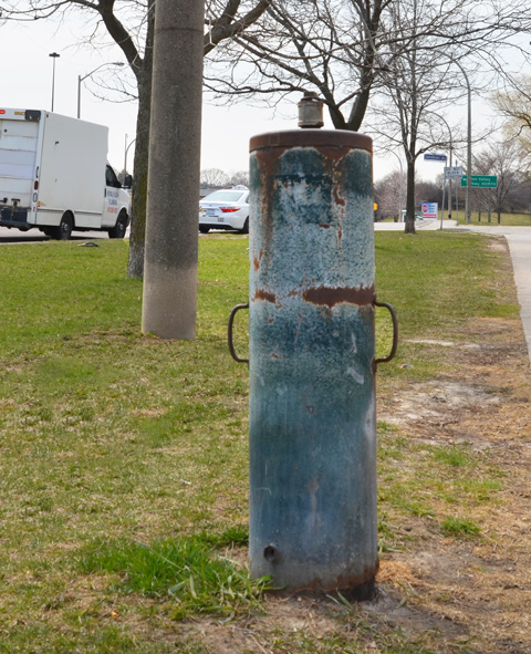 Small cylindrical metal object standing upright on the grass beside a sidewalk, rusty and old