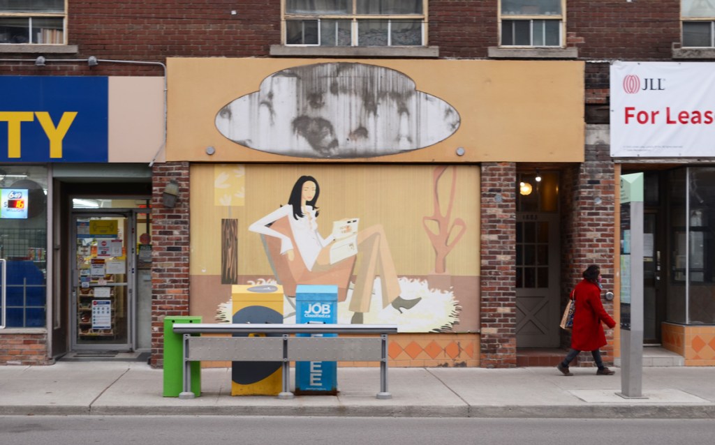 from across the street, mural of a woman on a sofa reading a book, mural, covers window of an empty store, a woman in a red coat is walking past, the store to the right is for lease, the store to the left is a variety store with a blue and yellow sign, 