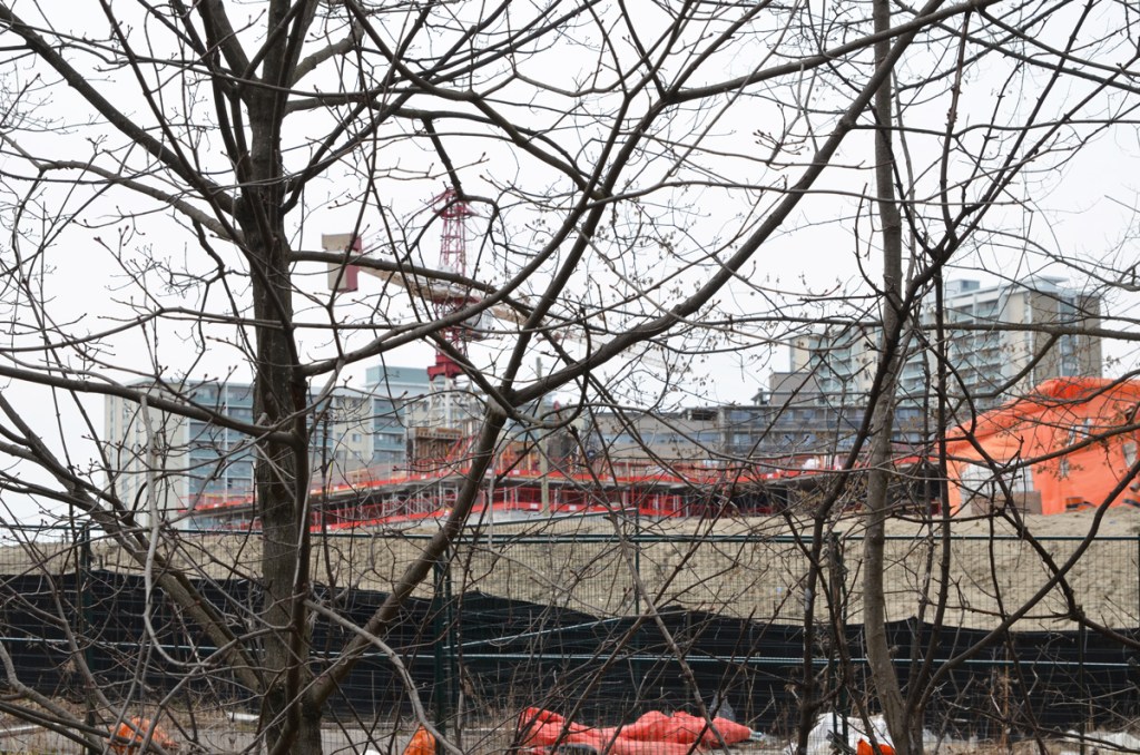 View from a park towards a construction site with first few floors of a new condo built. Taller apartment buildings in the background