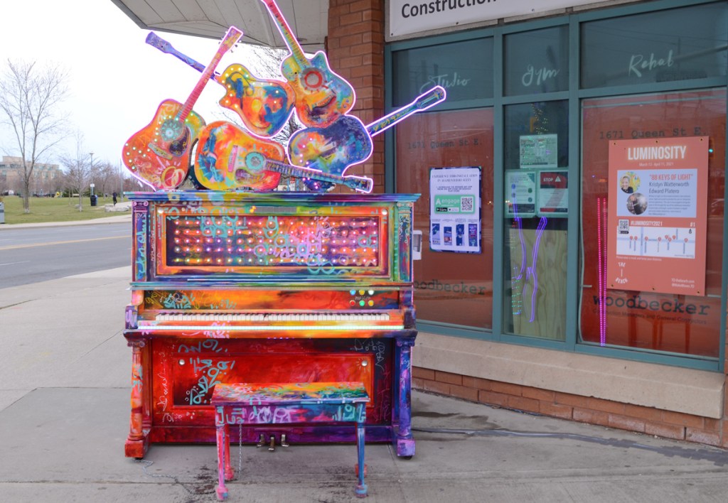 upright painted piano sits on the sidewalk, five guitars on top if it