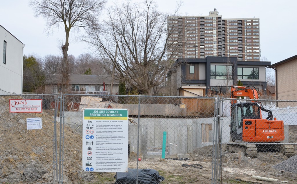 Construction site in residential area, house is mostly demolished, just footprint remains, fence around site, orange digger, backs of houses in next street, a tall apartment building in the background