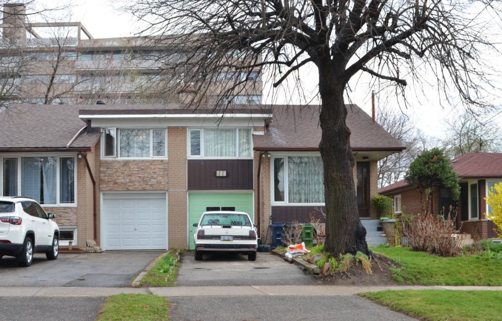 split level semi divided house with cars parked in the driveway, a large tree in front of the one on the right, apartment building behind 