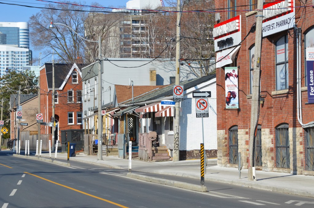 Looking west from Parliament street, the north side of Shuter Street including the Shuter St. Pharmacy, entrance to Poulett St, and some older small houses.  Taller, newer buildings in the background