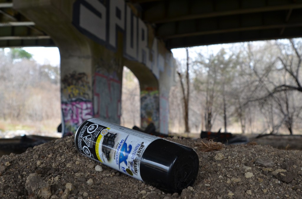 close up of a discarded can of spray paint lying on the ground.  Graffiti under a bridge is in the background but out of focus.