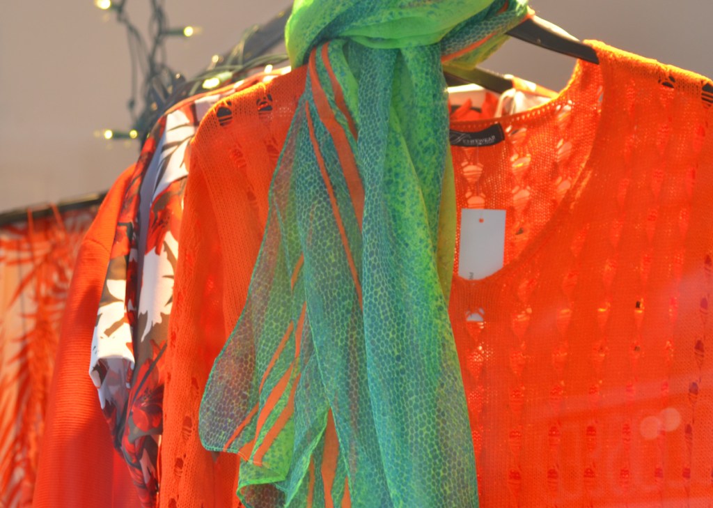 clothing store window with women's tops and blouses in orange hanging from a bar, a green and orange scarf around the neck of the orange knit top in front