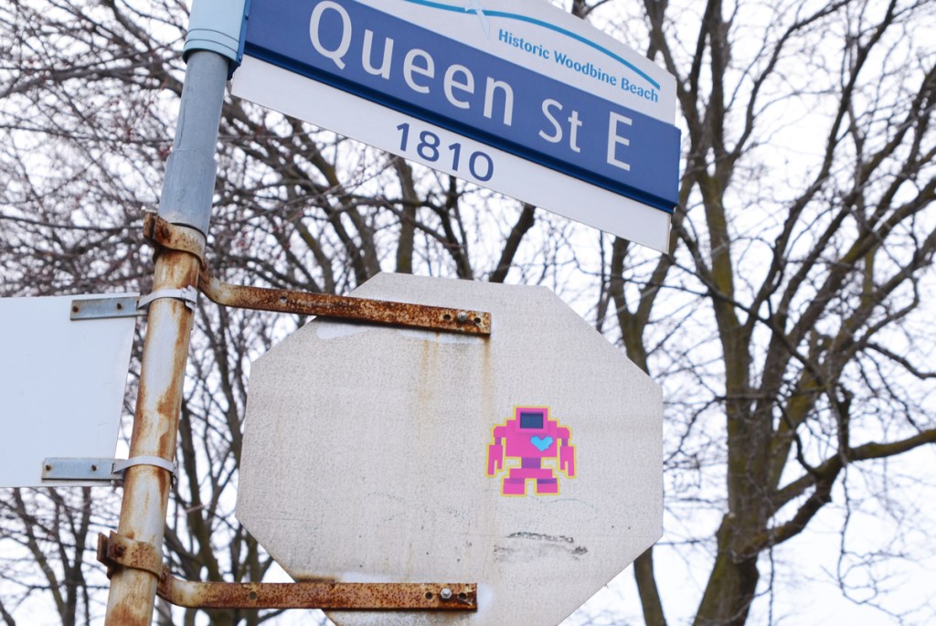 pink lovebot sticker, with a blue heart, on the back of a stop sign, historic woodbine beach street sign for Queen Street East as well 