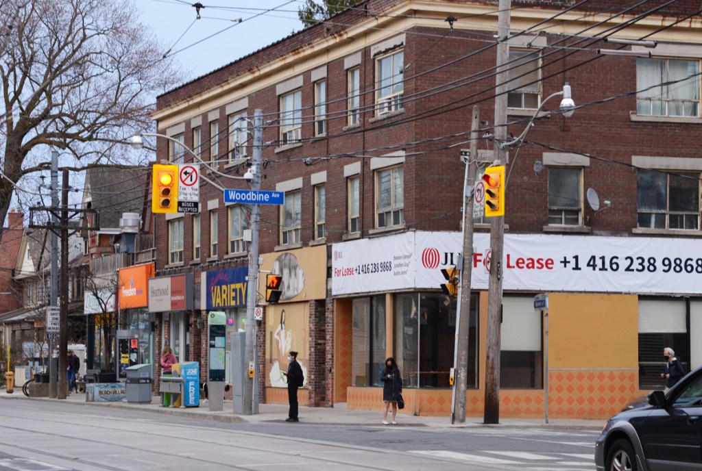 southeast corner of Woodbine and Queen St East, three storey brick building with stores on ground level, people waiting at traffic light, 