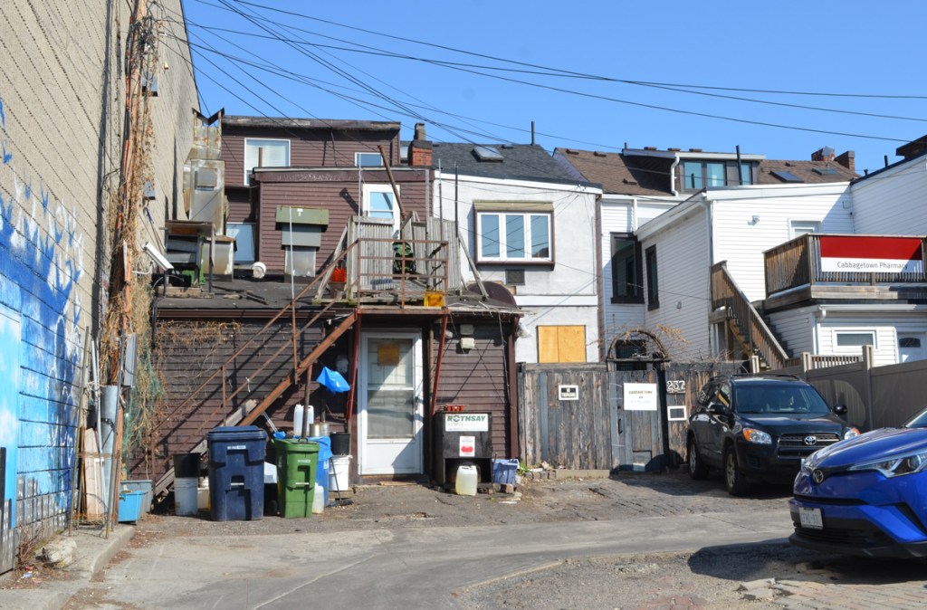 in an alley, view of back of houses with exterior staircases, fences, rubbish bins, parked cars, jumble of lines