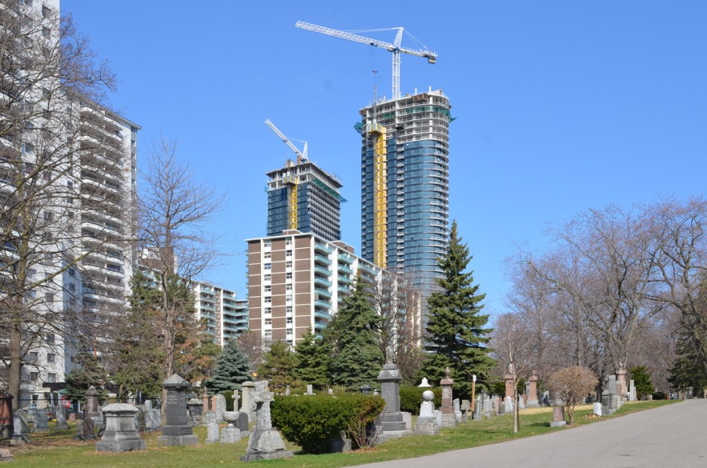 view from inside St. James cemetery, looking over some tombstones and monuments with large highrise of St. James town and newer condo development being built in the background, cranes, under construction, 
