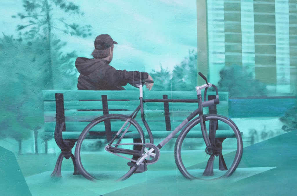 painting of a young man sitting on a bench, with his bicycle leaning against the back of the bench, an apartment building in the background, part of a mural featuring views of the east end of Toronto
