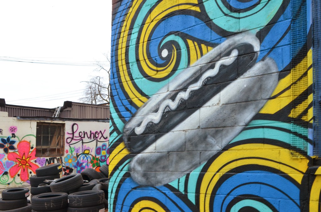 in the foreground, part of mural with blue, green, and yellow swirls.  A hot dog with wiggly line of mustard down the center of the wiener is on top of the swirls